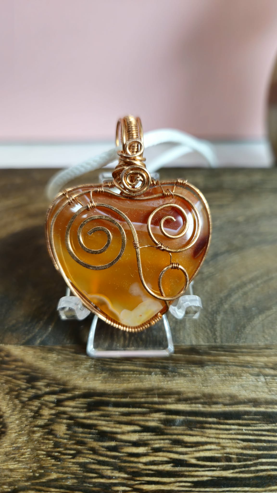 A heart-shaped Carnelian agate pendant wrapped in copper wire with spiral patterns, displayed on a clear stand over a wooden surface.