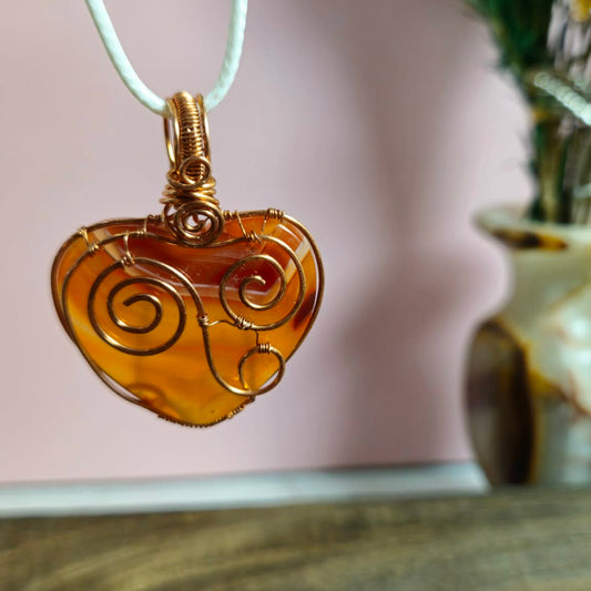 A Carnelian Agate heart-shaped pendant with a copper wire swirl design, hanging from a white cord against a soft pink background with a blurred wooden table and green plant in the foreground.