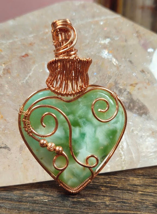 A heart-shaped green aventurine stone pendant wrapped in copper wire, resting on a wooden surface with a polished stone slab behind it.