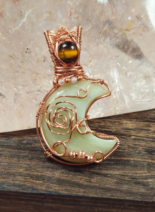 A handmade wire-wrapped pendant featuring a Green Jade crescent moon paired with a gold Tiger Eyer, a spiral rose motif, and a small copper & tourmaline beads. resting on a dark wooden surface with a crystal background.