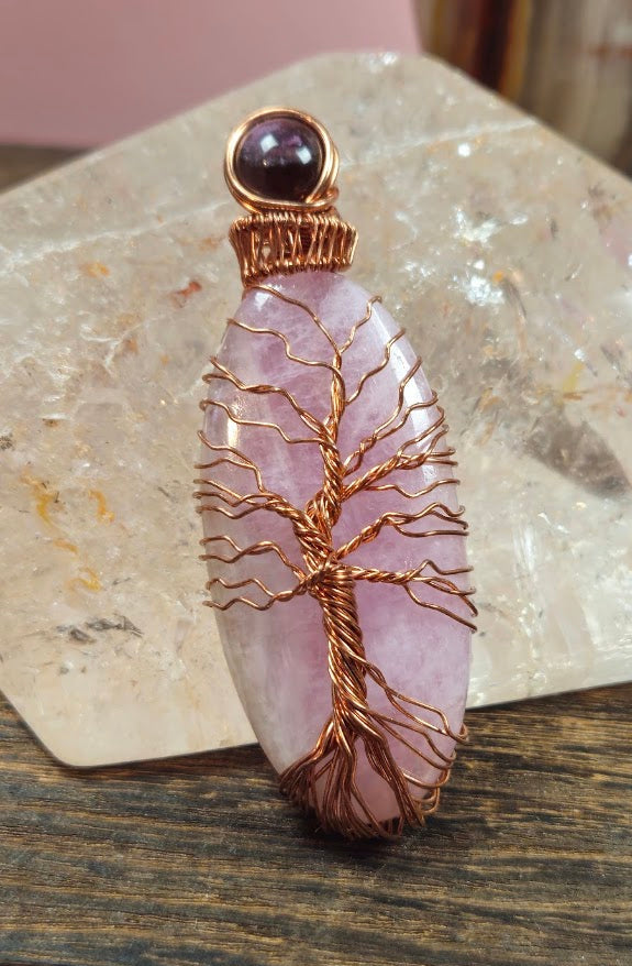 A handmade pendant featuring a pink Kunzite stone & smaller Amethyst stone with a copper wire tree of life design, displayed on a wooden surface in front of a clear quartz crystal.