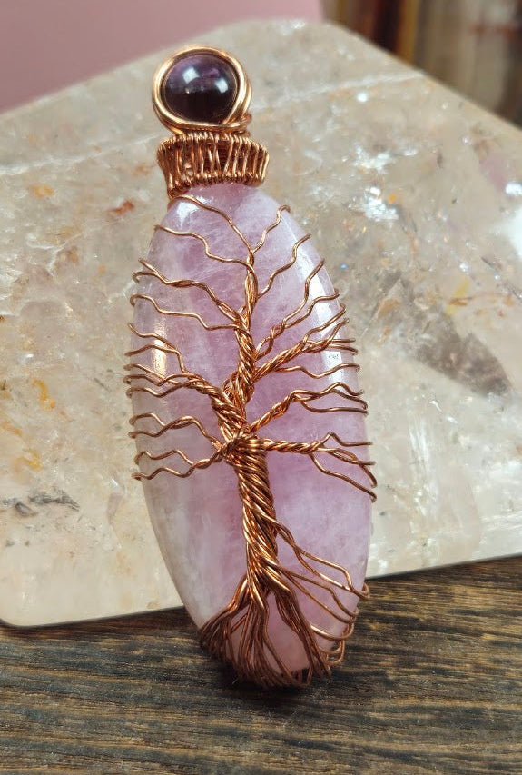 A handmade pendant featuring a pink Kunzite stone & smaller Amethyst stone with a copper wire tree of life design, displayed on a wooden surface in front of a clear quartz crystal.